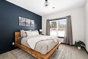 A bedroom with a bed, a window with curtains, a wall-mounted artwork, and a potted plant.