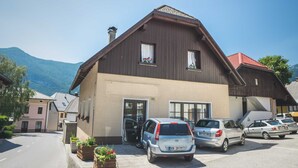 A two-story building with a brown facade, two visible windows with curtains, and a red roof. There are several cars parked in front, and a mountain is visible in the background.