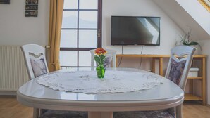 A dining area with a round table, chairs, a vase with a flower, a television, and a window with curtains.