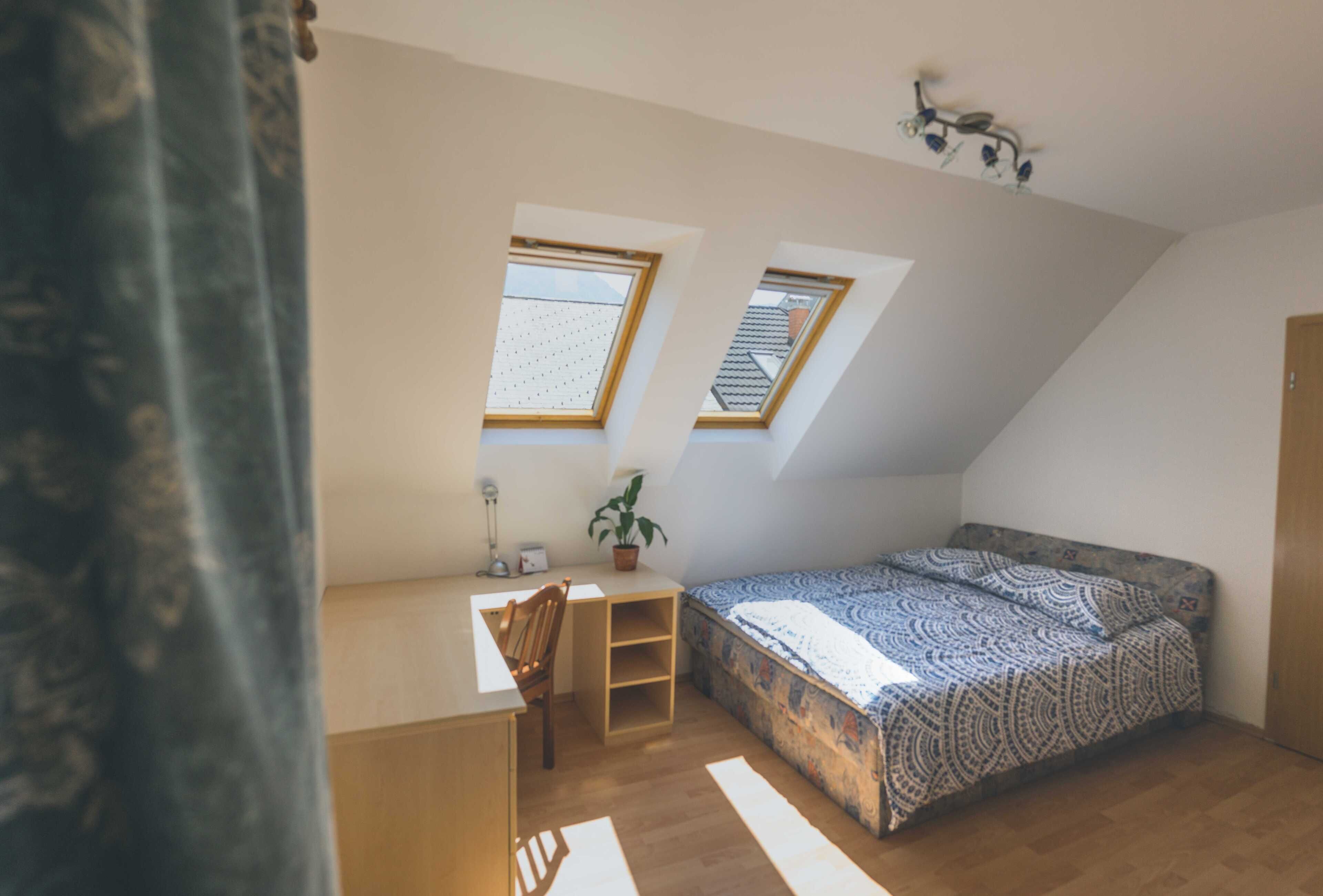 A small, cozy attic bedroom with a sloped ceiling, two skylights, a desk, a chair, and a bed with patterned bedding.