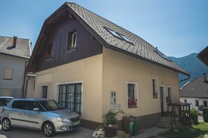 Un bâtiment à deux étages, doté d’un toit en pente, avec une voiture garée devant et un petit jardin agrémenté de plantes en pot.