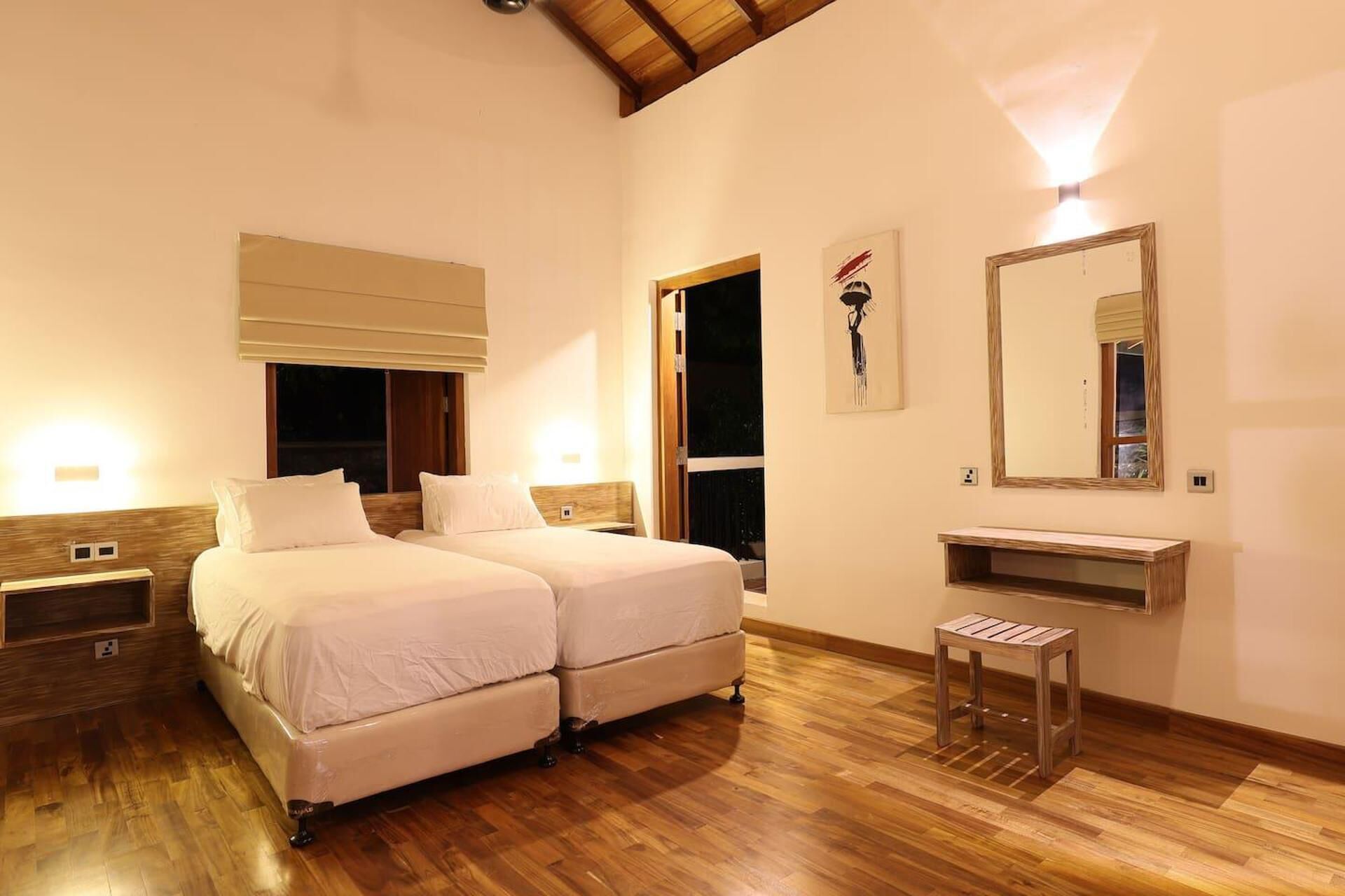 A hotel room with two beds, a wooden floor, a nightstand, a bedside table, a mirror, and a window with a view.