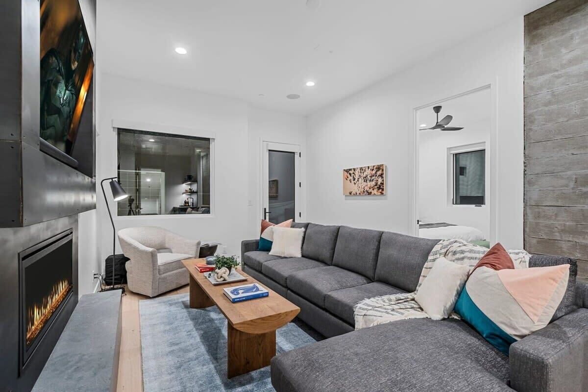 A modern living room with a fireplace, grey sofas, a wooden coffee table, and a large window.