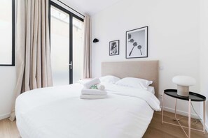 A neatly made bed with white linens and a rolled towel, a small bedside table with a lamp, and two framed pictures on the wall. - Appartement 2 chambres, idéal famille et amis (Paris)