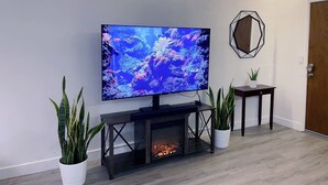A modern living room with a large flat-screen TV mounted on a black console table, a fireplace, and two potted plants.