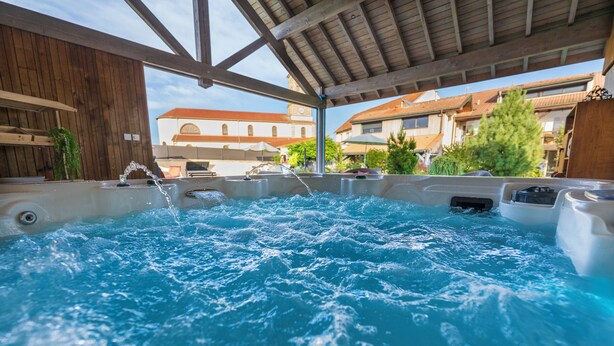 A hot tub with water flowing, surrounded by wooden structures and a view of residential buildings. - La Chambre Enchantée (La Baffe)