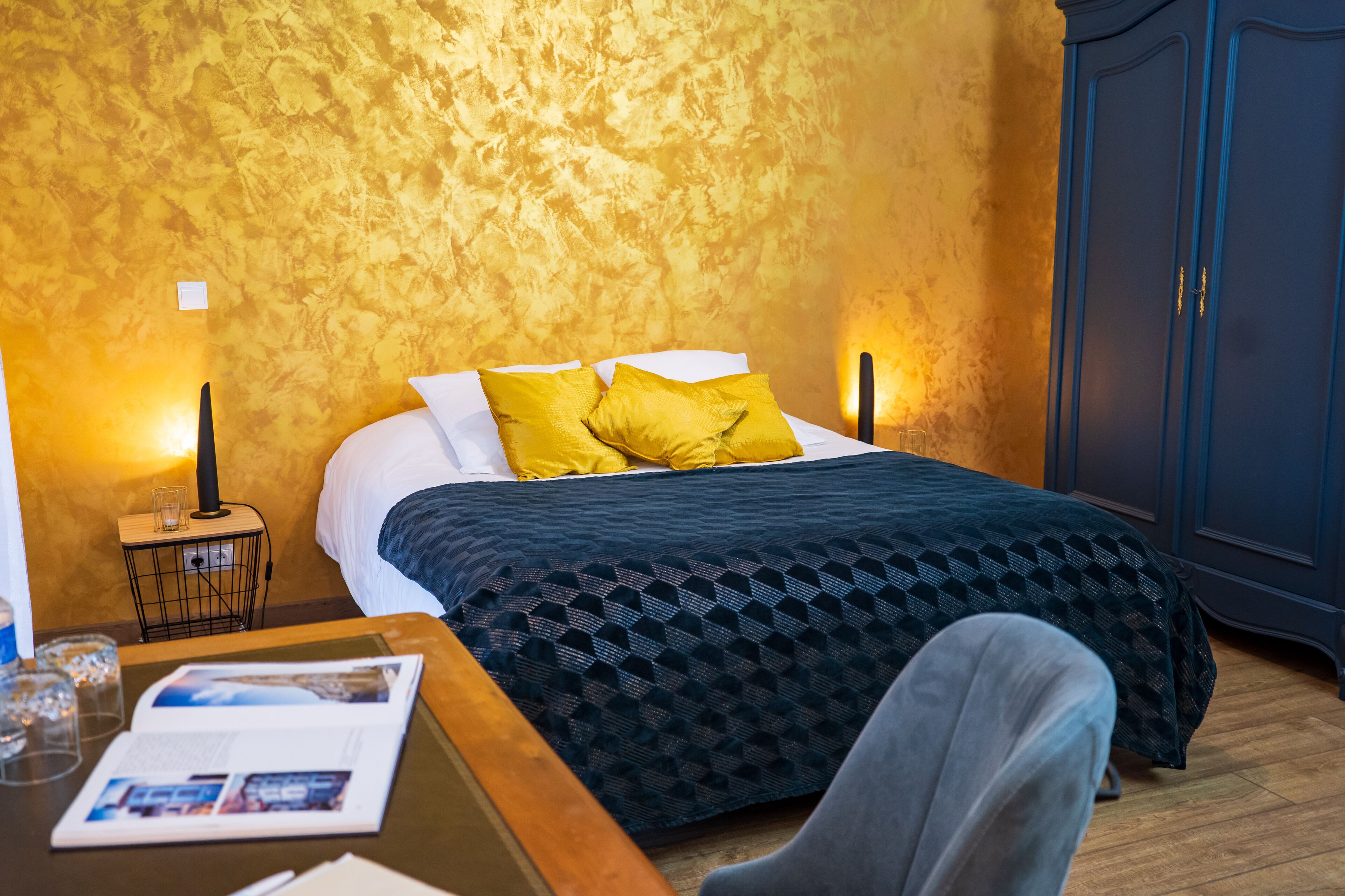 Una habitación de hotel con una cama, una mesita de noche con lámpara, una mesa de madera con una revista y un armario.