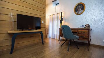 Une pièce avec un plancher en bois, un bureau avec une chaise, une télévision, un tableau encadré au mur et une fenêtre avec des rideaux.