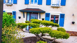 A building with a brick facade, blue shutters, and a small garden with a bonsai tree. - La Chambre Enchantée (La Baffe)