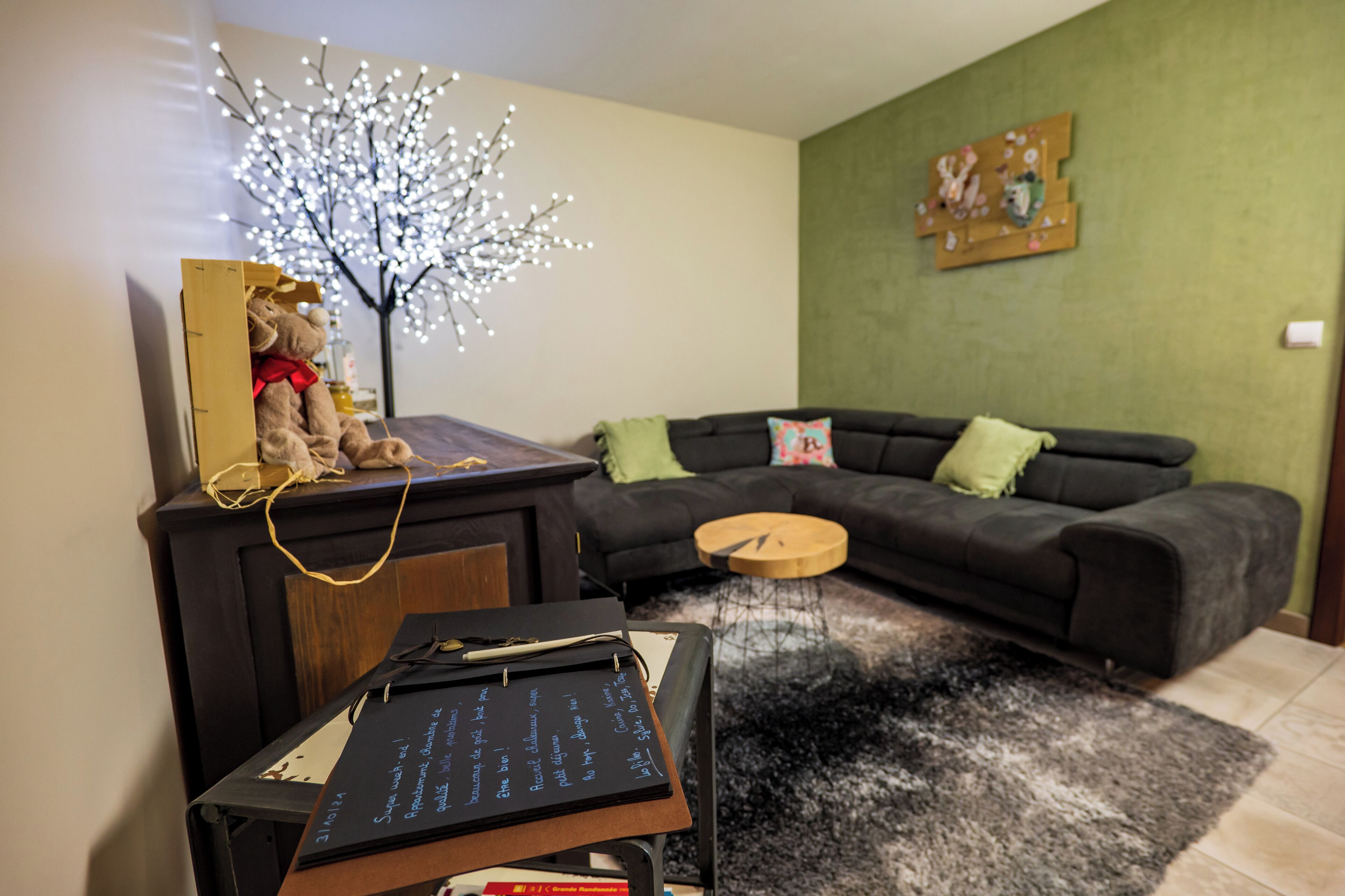 Un salon avec un canapé de couleur sombre, une petite table d’appoint en bois, un tableau noir avec des inscriptions, un arbre décoratif illuminé et un mur vert orné d’un tableau.