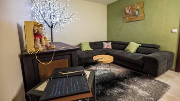 Un salon avec un canapé de couleur sombre, une petite table d’appoint en bois, un tableau noir avec des inscriptions, un arbre décoratif illuminé et un mur vert orné d’un tableau.