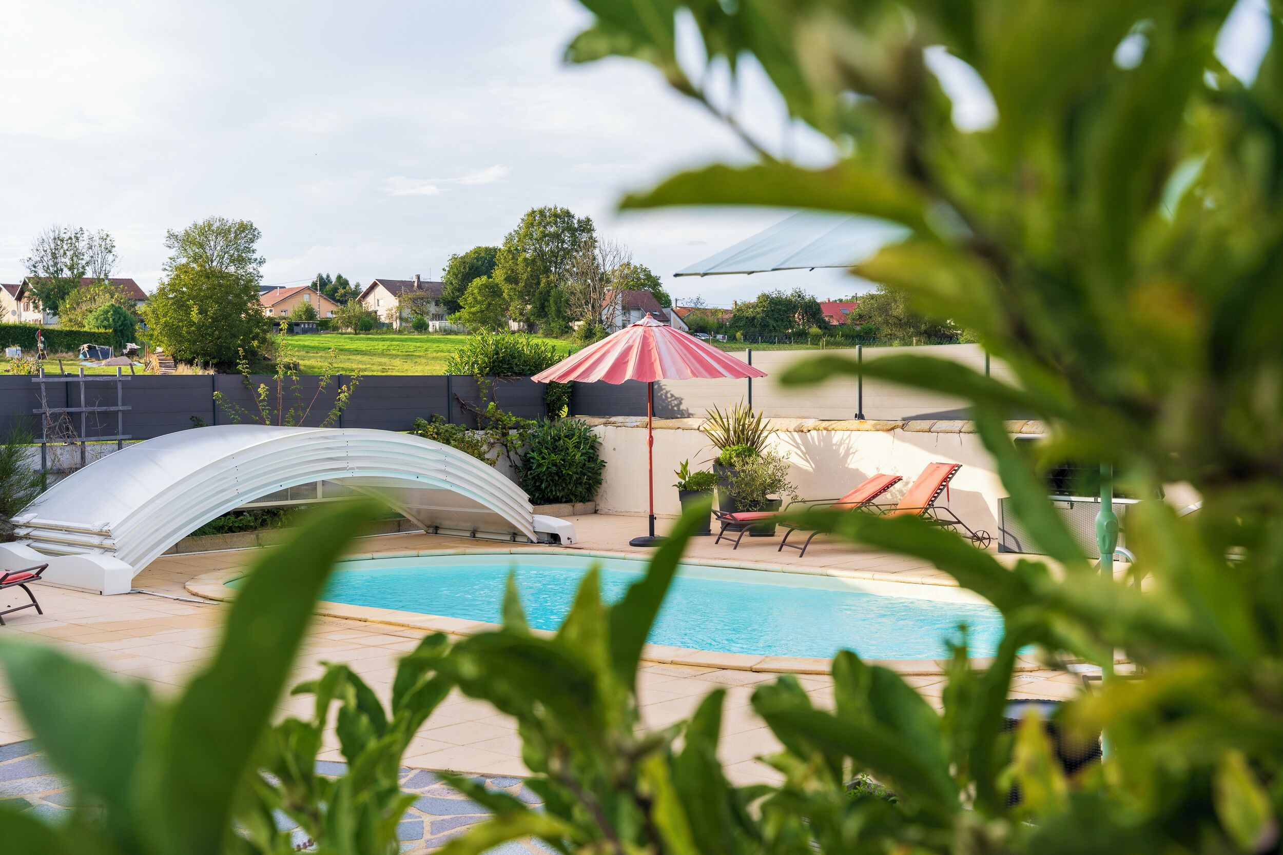 A swimming pool with a white canopy and red umbrella, surrounded by lounge chairs and potted plants.