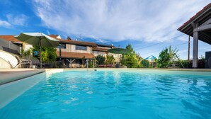 A swimming pool with a clear blue water surface, surrounded by a covered patio area with umbrellas and outdoor furniture. - La Chambre Enchantée (La Baffe)