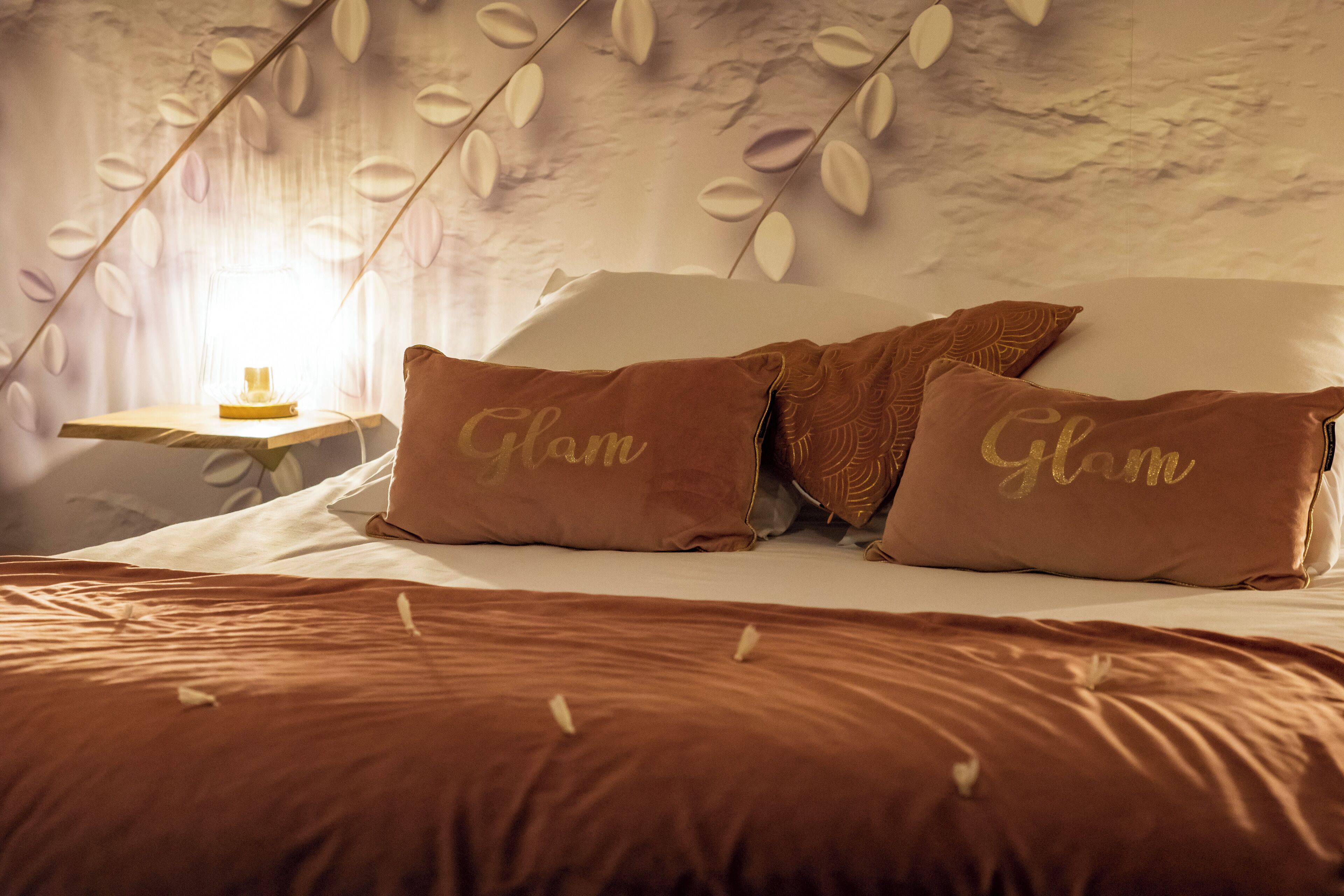 A bedroom with a bed, two pillows with "Glam" text, a nightstand with a lamp, and a wall with leaf patterns.