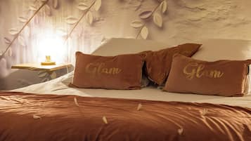 A bedroom with a bed, two pillows with "Glam" text, a nightstand with a lamp, and a wall with leaf patterns.