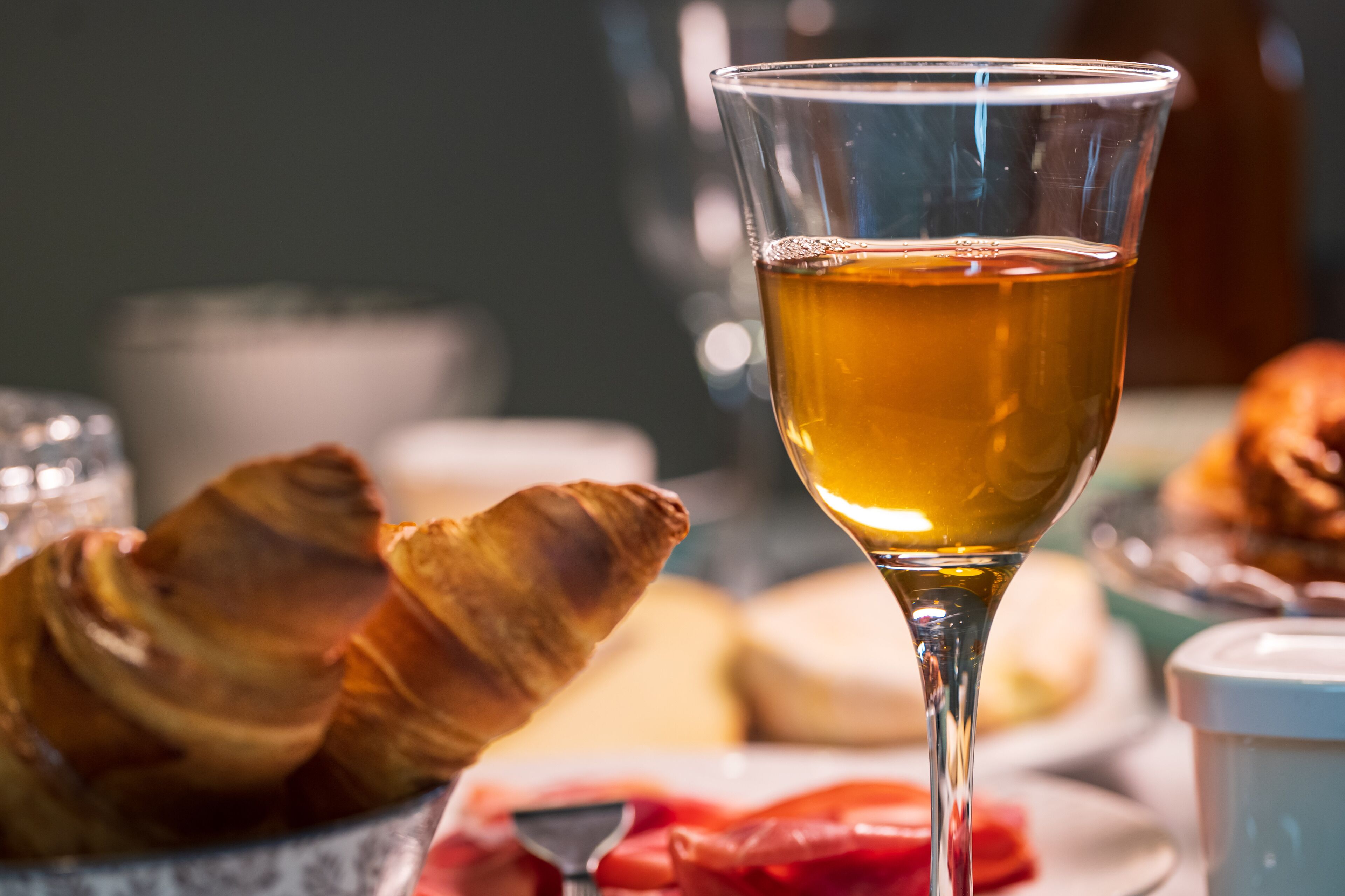 Um copo de líquido âmbar, possivelmente chá, com croissants e um prato de comida ao fundo.