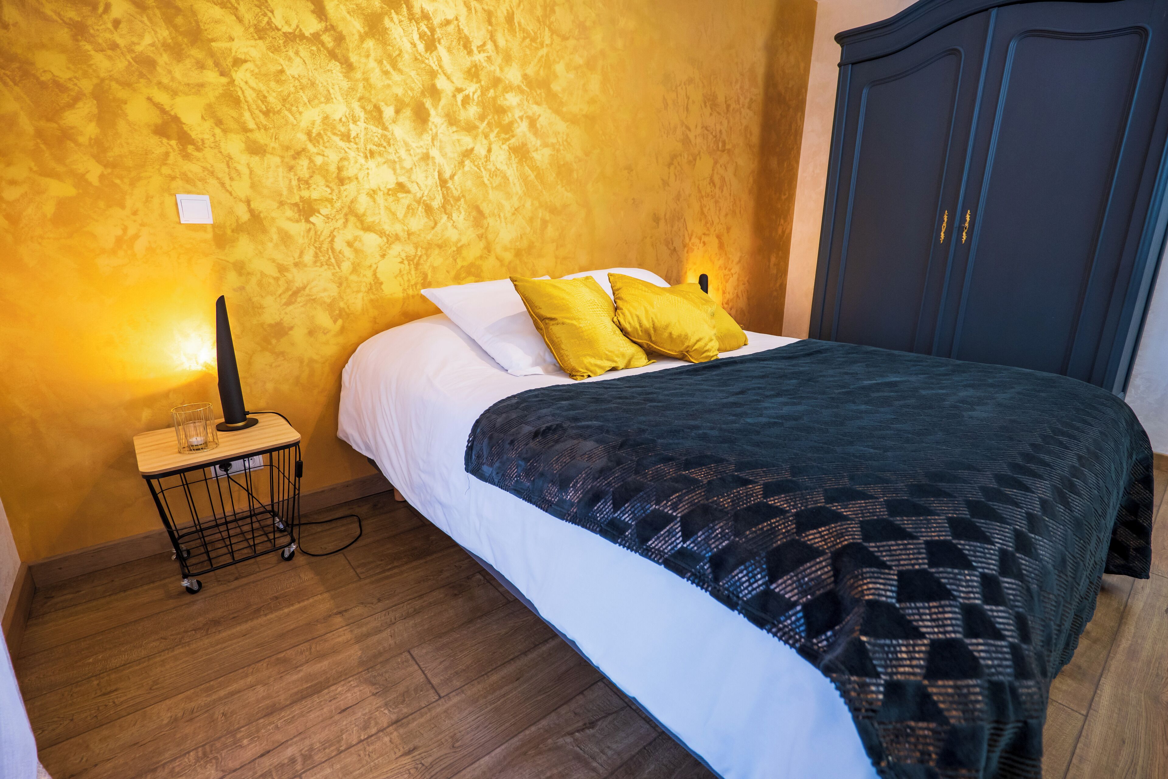 A bedroom with a bed, wooden floor, a nightstand with a lamp, and a wardrobe.