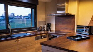 A modern kitchen with wooden cabinets, a built-in oven, a coffee maker, and a window overlooking a grassy area. - La Chambre Enchantée (La Baffe)