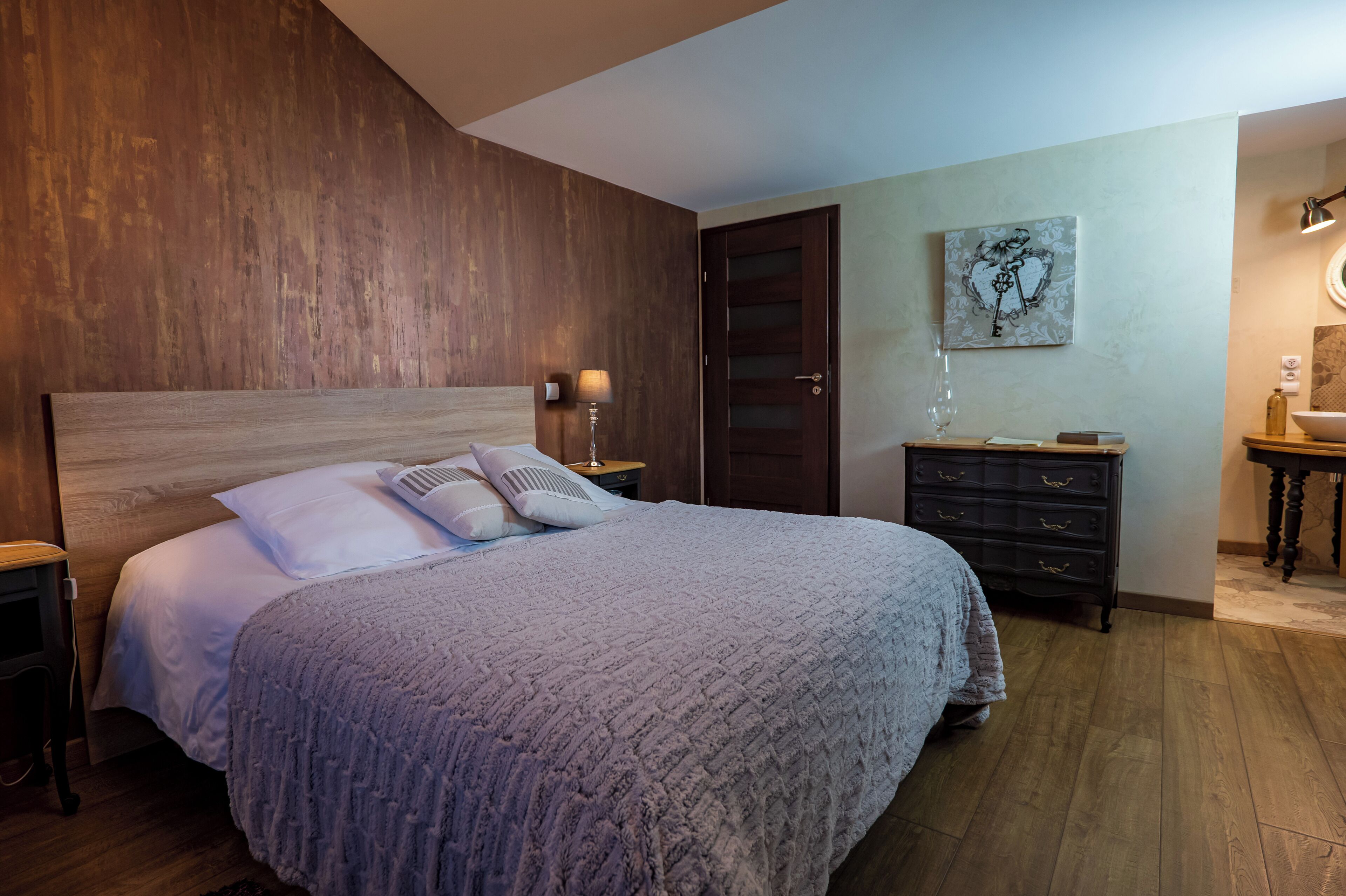 Een slaapkamer met een houten hoofdbord, een bed met een grijze deken, een nachtkastje met een lamp, een dressoir en een aan de muur hangend kunstwerk.