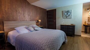 Een slaapkamer met een houten hoofdbord, een bed met een grijze deken, een nachtkastje met een lamp, een dressoir en een aan de muur hangend kunstwerk.