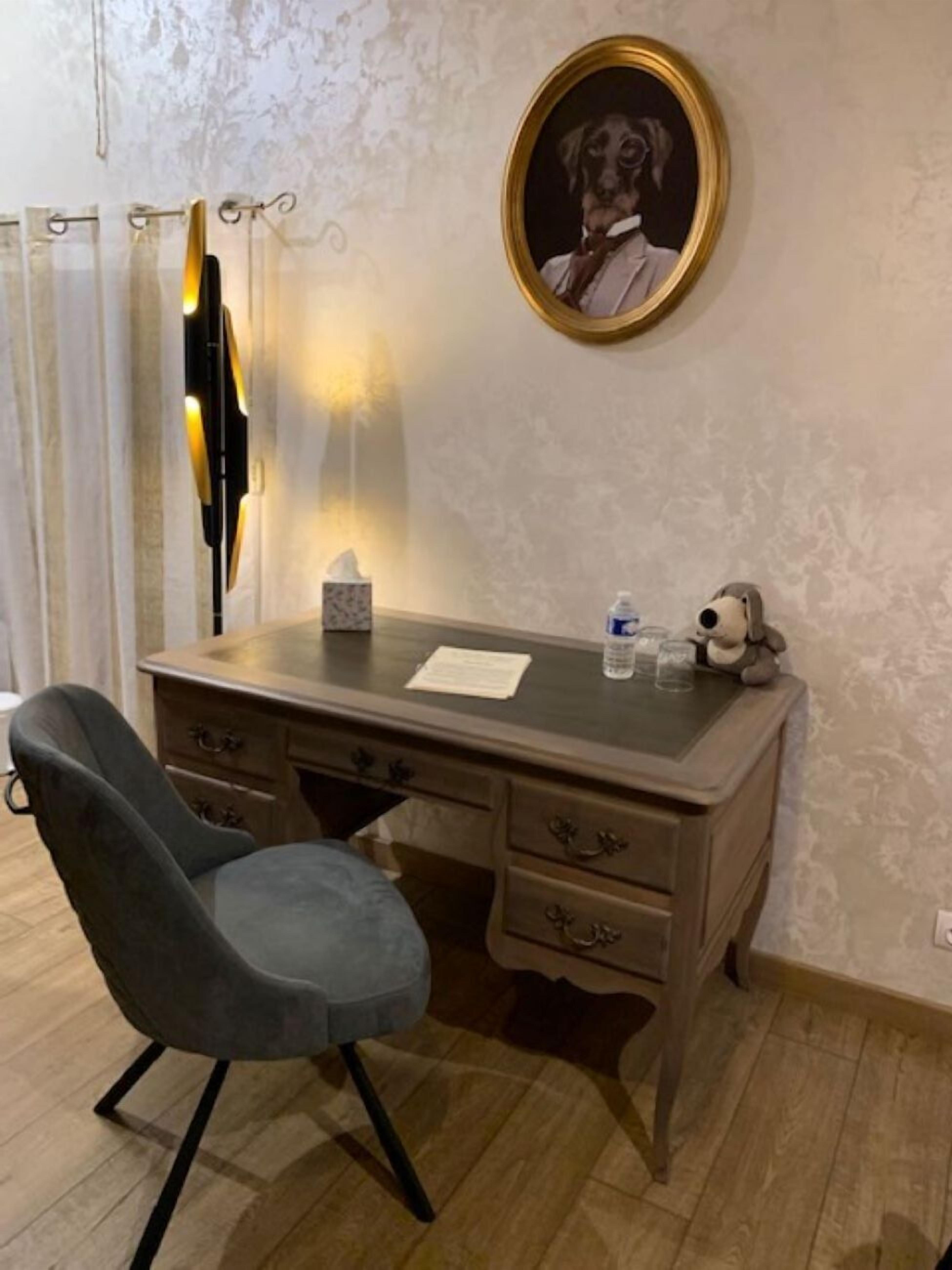 A room with a wooden desk, a chair, a framed portrait on the wall, a lamp, and a small table with a bottle and glasses.