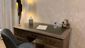 A room with a wooden desk, a chair, a framed portrait on the wall, a lamp, and a small table with a bottle and glasses.