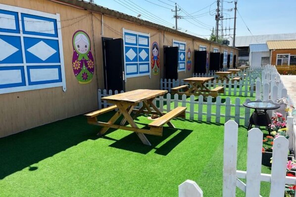 Une rangée de tables et de bancs de pique-nique en bois, une clôture blanche à piquets et un bâtiment orné de peintures murales colorées.