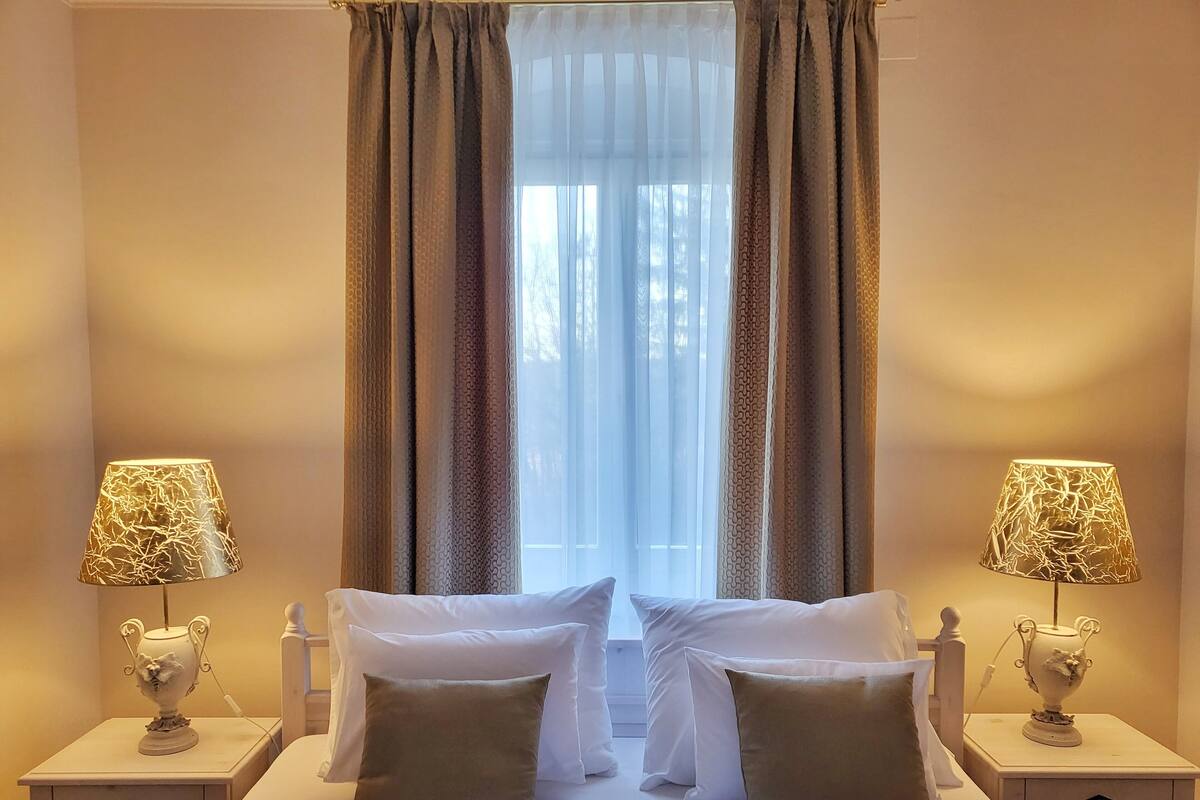 A hotel room with two beds, a large window with sheer curtains, and two bedside lamps with decorative bases.