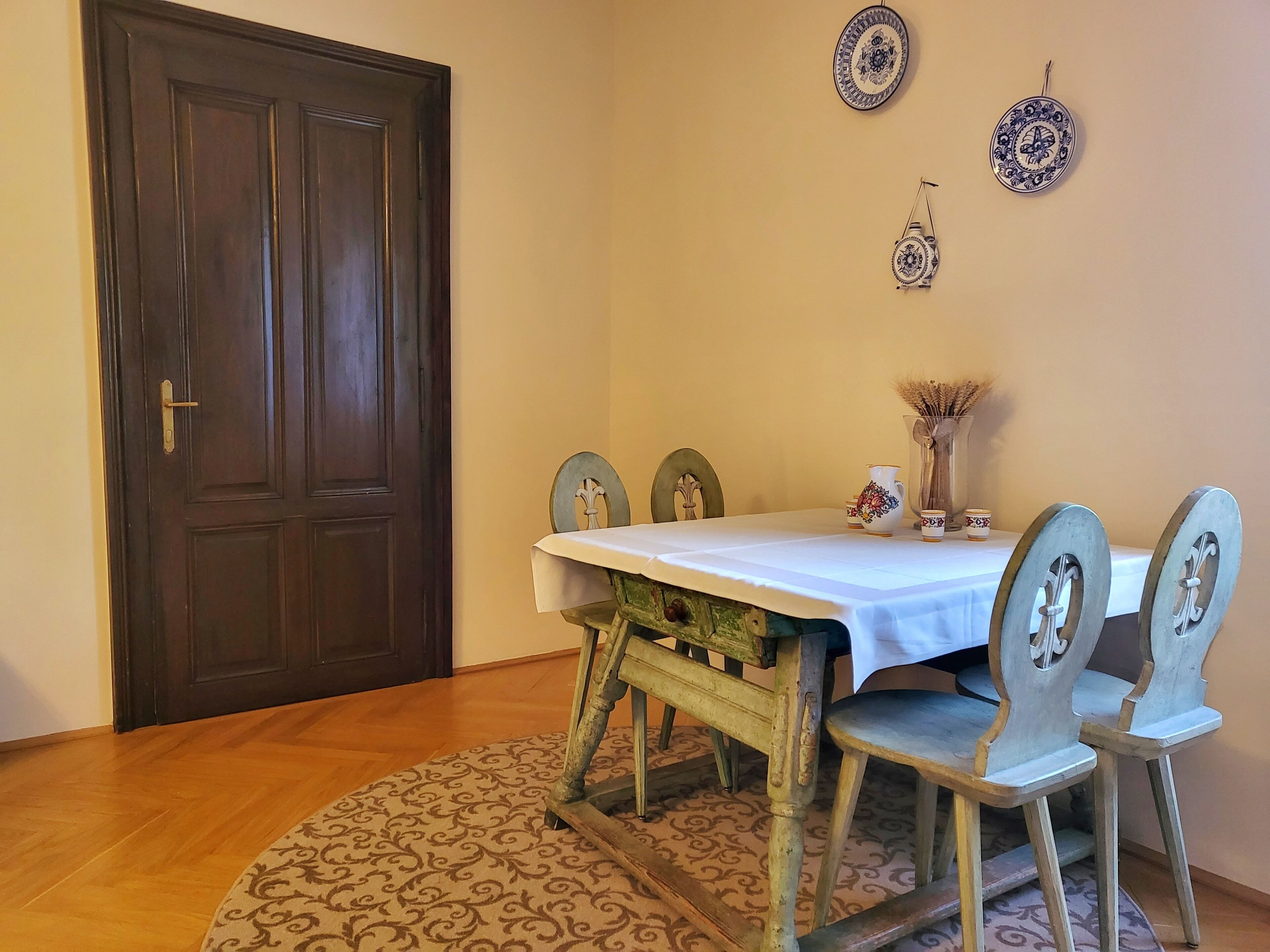 A dining area with a wooden table, four chairs, a patterned rug, and decorative plates on the wall.