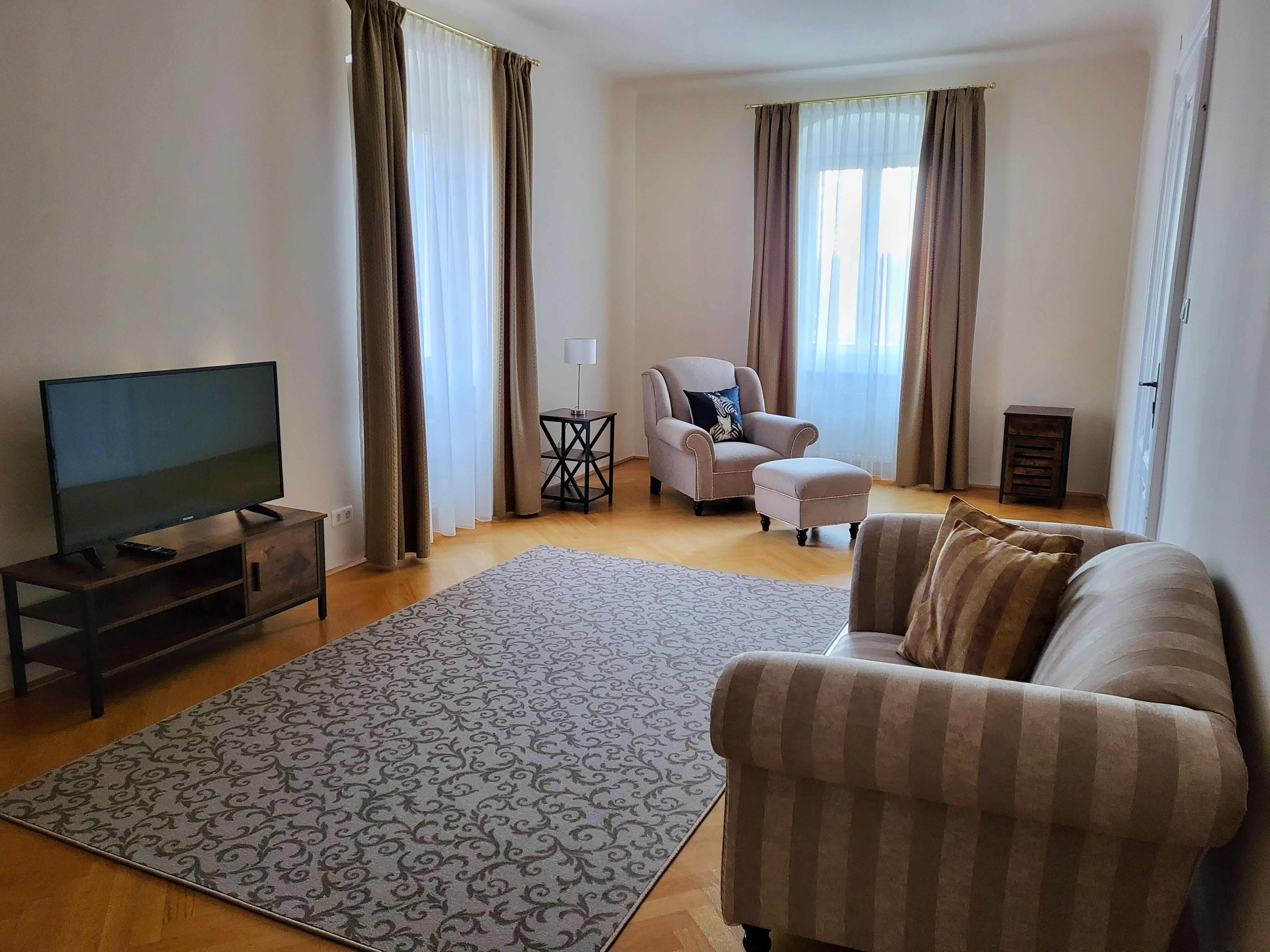 A living room with a flat-screen TV on a wooden stand, a beige sofa, a patterned rug, a small side table with a lamp, and a chair with a cushion.