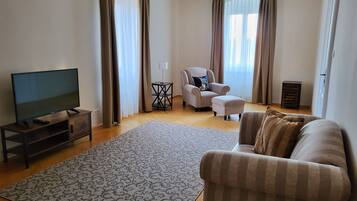 A living room with a flat-screen TV on a wooden stand, a beige sofa, a patterned rug, a small side table with a lamp, and a chair with a cushion.