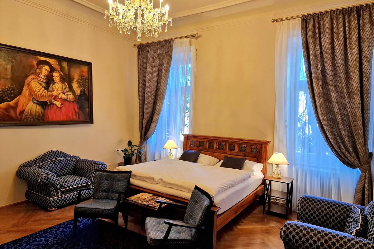 A hotel room with a large bed, two armchairs, a wooden headboard, a chandelier, and a painting on the wall.