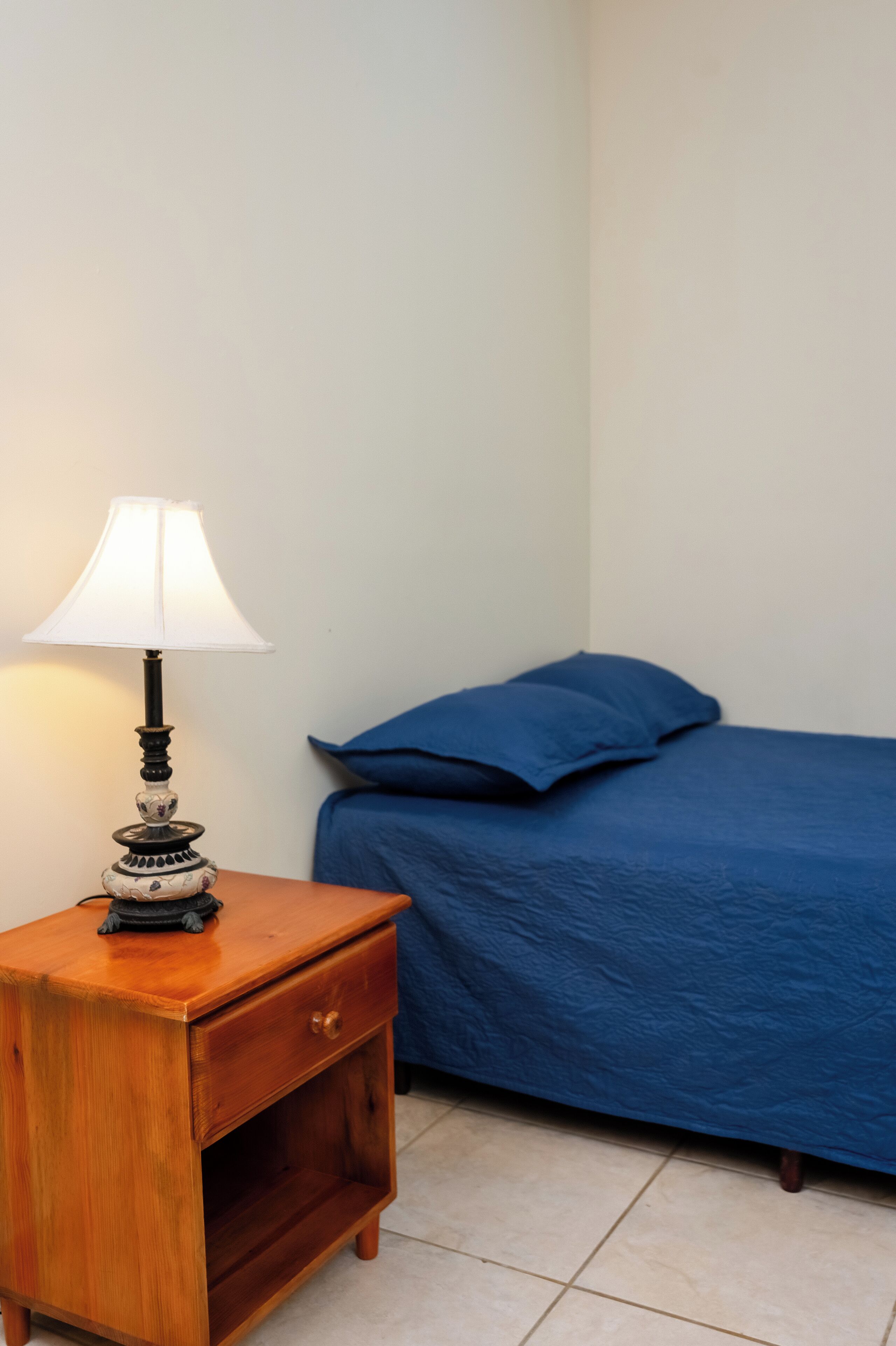 Une petite table de chevet en bois avec une lampe, un lit bleu avec des oreillers et un mur blanc uni.