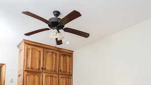 Un dormitorio con un armario de madera, un ventilador de techo, una mesita de noche con lámpara y una cama con cubrecama azul.