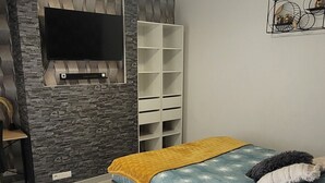 Ett sovrum med en säng, en tv som är monterad på en vägg av sten, en vit hylla och ett nattduksbord i trä.