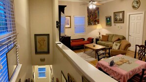 Uma sala de estar com sofá, mesa de centro e mesa de jantar com toalha de mesa rosa. Há quadros nas paredes e um ventilador de teto.