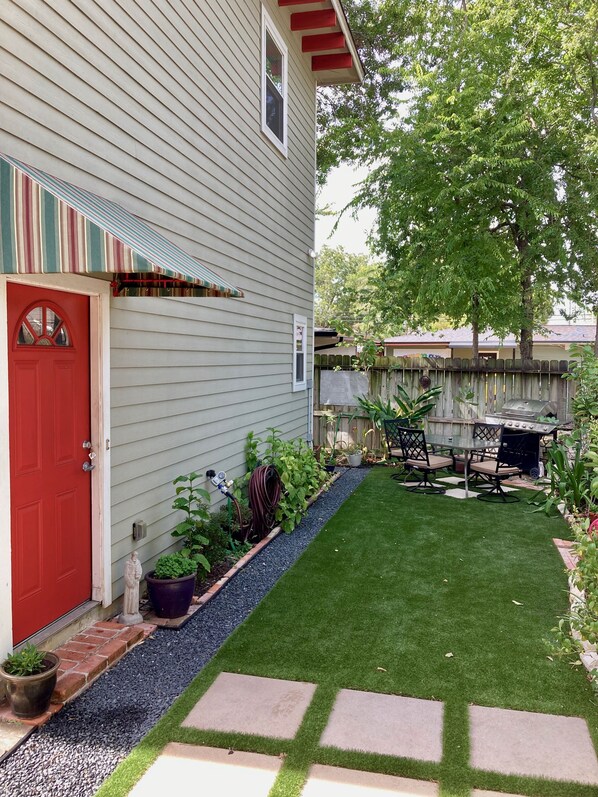 A house with a red door, a small garden with a lawn and stepping stones, and a barbecue area.
