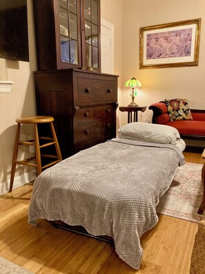 A bedroom with a bed, a wooden dresser, a stool, a lamp, and a framed picture on the wall.