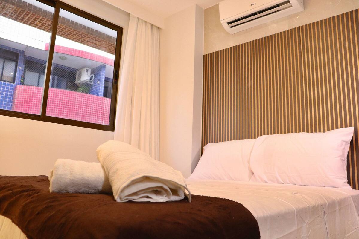Habitación de hotel con cama, almohadas blancas, una manta marrón y ventana con vista a un edificio.