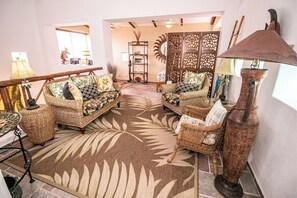 Een woonkamer met rieten meubilair, een kamerkleed met dessin en een decoratief paneel aan de muur.