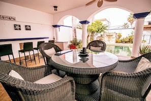 A round table with wicker chairs, a glass top, and a view of a swimming pool and greenery outside.