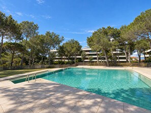 A swimming pool with a wooden deck, surrounded by trees and a fence, with a building in the background. - Apartamento Nuevo, Equipado y Decorado con Estilo (Palamós)