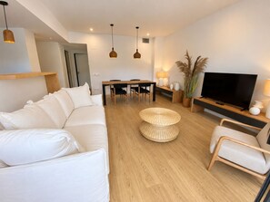 A modern living room with a white sofa, a dining table with chairs, a TV, and a large plant. - Apartamento Nuevo, Equipado y Decorado con Estilo (Palamós)
