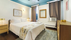 A hotel room with a bed, bedside lamps, a ceiling fan, a window with curtains, an air conditioning unit, and a wooden dresser. - Newly Updated 3BR Furnished Home Close to Path, Lightrail and Pets Welcome (Jersey City)