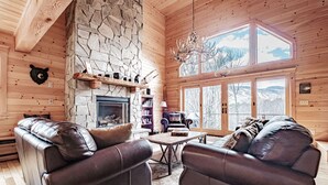 A cozy cabin interior with a stone fireplace, leather sofas, and a large window offering a mountain view. - Spring & Summer Specials (Newry)