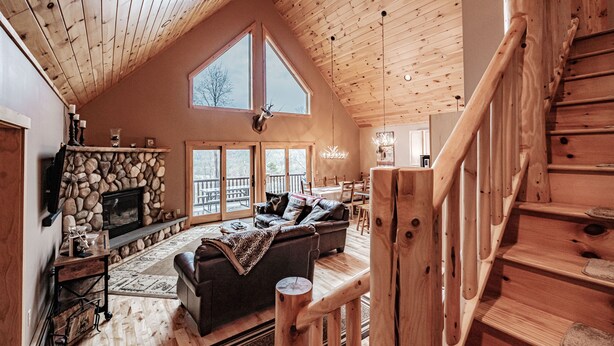 A spacious living room with a fireplace, a stone wall, a large window, and a wooden staircase.
