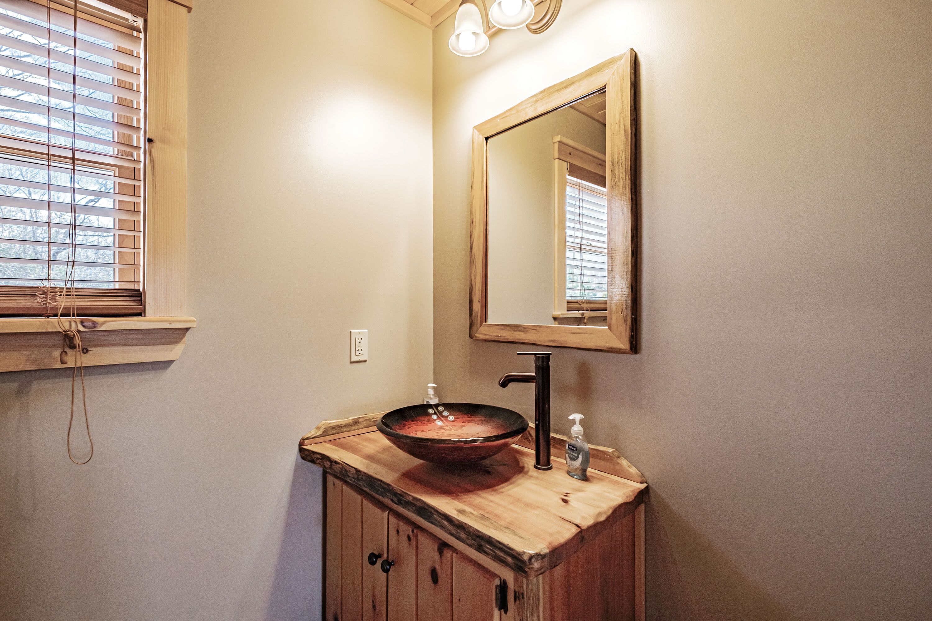 Un baño con tocador de madera, un lavabo de cerámica, un espejo y una ventana con persianas.