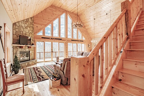 A spacious living room with a stone fireplace, large windows, a wooden staircase, and a cozy seating area.