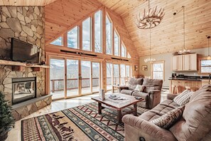 A spacious living room with a stone fireplace, large windows, a cozy sofa, and a wooden ceiling.
