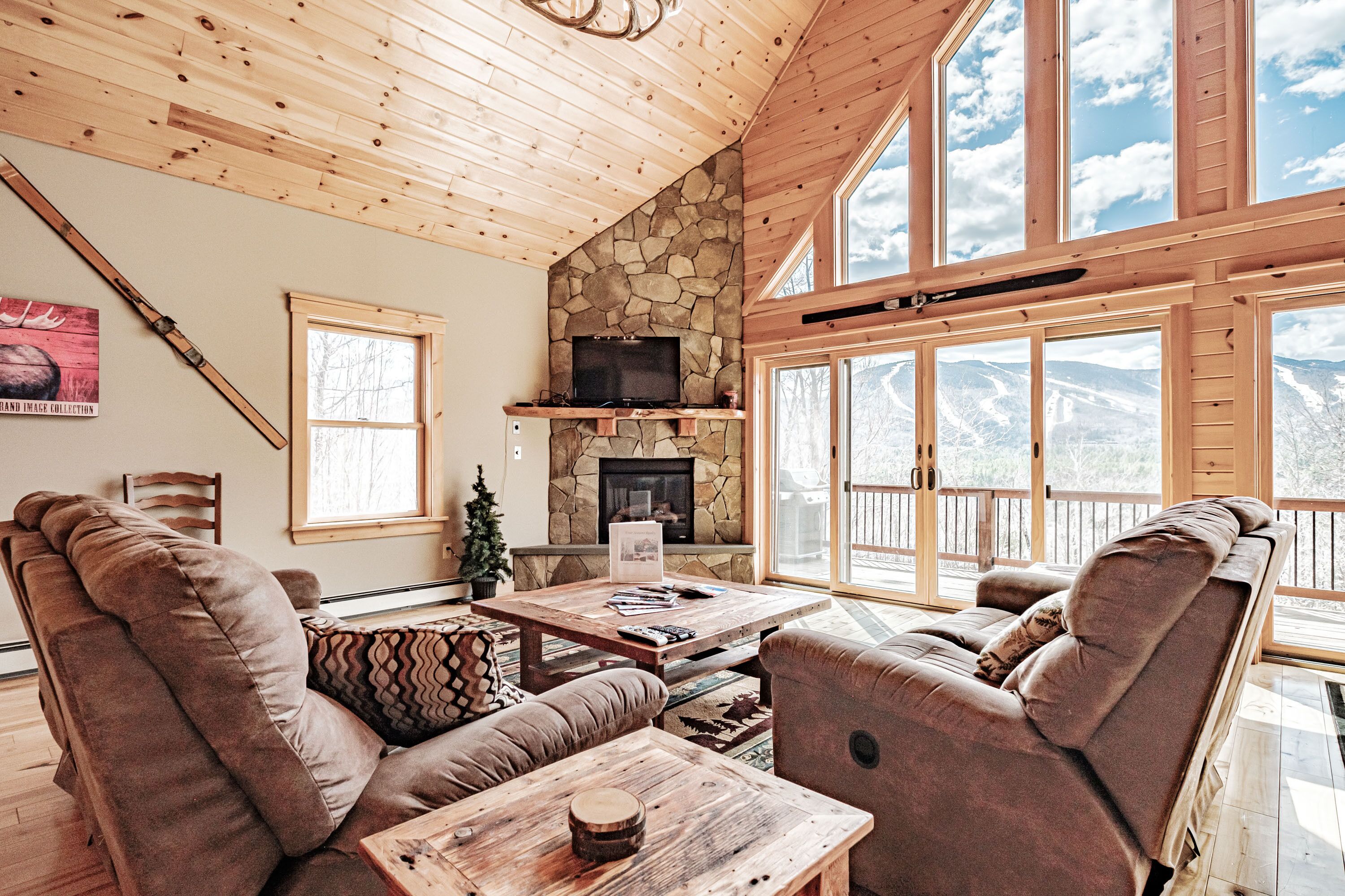 A cozy living room with a stone fireplace, wooden ceiling, and large windows overlooking a snowy landscape.